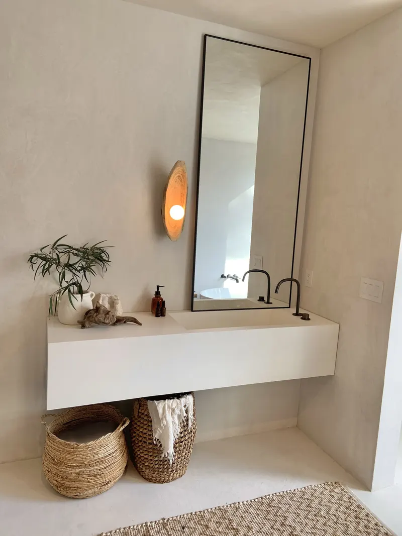 A floating vanity with a tall mirror, a warm wall sconce and woven baskets beneath